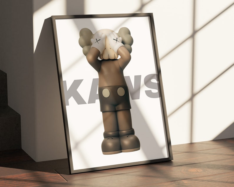 May include: A framed print of a brown and white Kaws figure with its hands covering its eyes. The figure is standing on a white background and the word "KAWS" is printed in gray on the right side of the image.