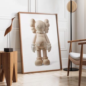 May include: A framed print of a beige KAWS Companion figure with white gloves and shoes. The artwork is displayed in a room with a wooden side table, a wooden bird sculpture, and a modern chair. The room has white walls and a wooden floor.