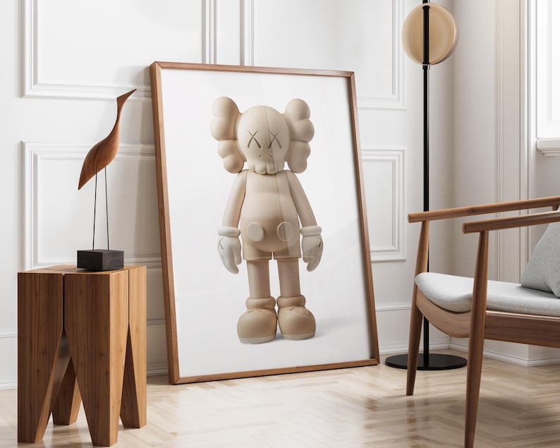 May include: A framed print of a beige KAWS Companion figure with white gloves and shoes. The artwork is displayed in a room with a wooden side table, a wooden bird sculpture, and a modern chair. The room has white walls and a wooden floor.