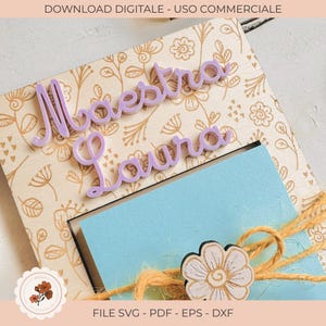 Pode incluir: Uma moldura de madeira com um design floral e as palavras "Maestra Laura" em escrita lavanda. Um cartão azul claro é visível, amarrado com barbante e um detalhe de flor de madeira. A imagem inclui o texto "DOWNLOAD DIGITALE - USO COMMERCIALE" e "FILE SVG - PDF - EPS - DXF".
