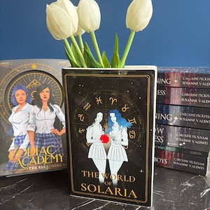 Puede incluir: Un jarrón negro y dorado con forma de libro con el texto "THE WORLD OF SOLARIA" y un diseño de zodíaco, que contiene tulipanes blancos. Un libro titulado "ZODIAC ACADEMY" es visible en el fondo, junto con otros libros.