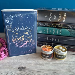 Könnte beinhalten: Ein blaues Buch mit dem Titel "Velaris City of Starlight" und einem goldenen Bergketten-Design. Das Buch liegt neben einem Stapel von fünf Büchern mit dem Titel "A Court of Silver Flames" und anderen Titeln der Reihe. Zwei kleine Kerzen mit den Namen "Lucien" und "Morrigan" stehen vor den Büchern.