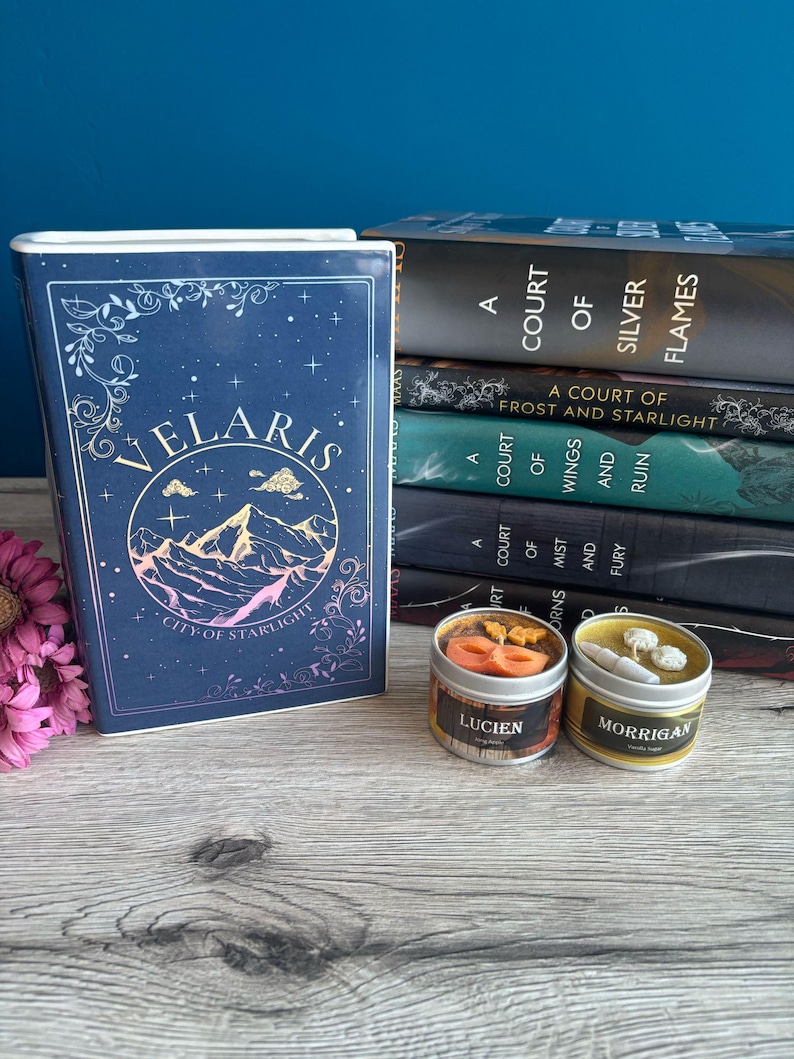 May include: A blue book with the title "Velaris City of Starlight" and a gold mountain range design.  The book is next to a stack of five books with the title "A Court of Silver Flames" and other titles in the series.  Two small candles with the names "Lucien" and "Morrigan" are in front of the books.