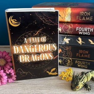 May include: A black book with gold lettering that reads "A Tale of Dangerous Dragons". The book is surrounded by a gold and black design. The book is sitting on a wooden table with two small golden dragon figurines.
