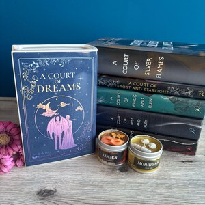 May include: A stack of seven fantasy novels with the title "A Court of Silver Flames" on top. The books are all blue and green with gold lettering. There are two candles in front of the books, one with the word "Lucien" and the other with the word "Morrigan".