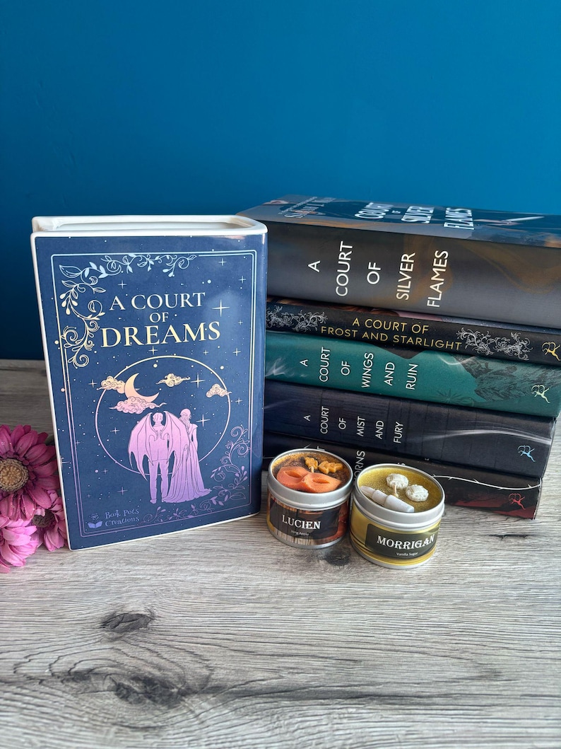 May include: A stack of seven fantasy novels with the title "A Court of Silver Flames" on top. The books are all blue and green with gold lettering. There are two candles in front of the books, one with the word "Lucien" and the other with the word "Morrigan".