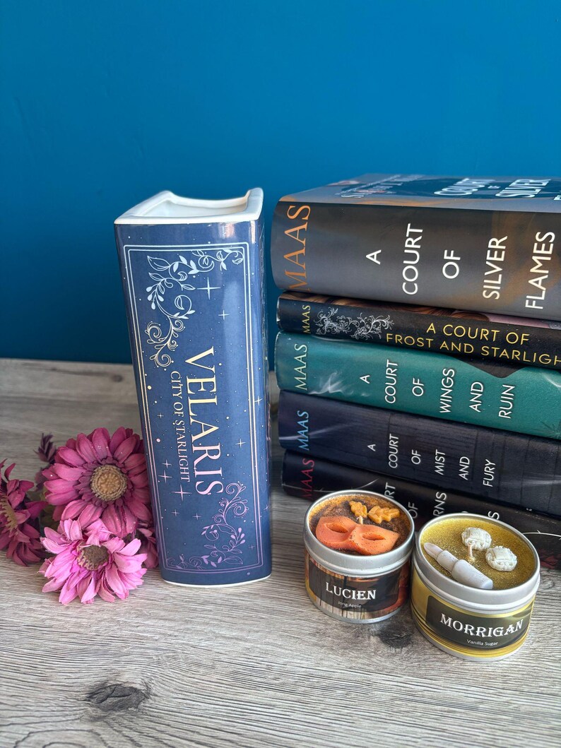 May include: A stack of six fantasy novels with the title "A Court of Silver Flames" on top. The books are by author Sarah J. Maas. A blue ceramic bookend with the words "Velaris City of Starlight" is in the foreground. Two small candles with the names "Lucien" and "Morrigan" are in the foreground.