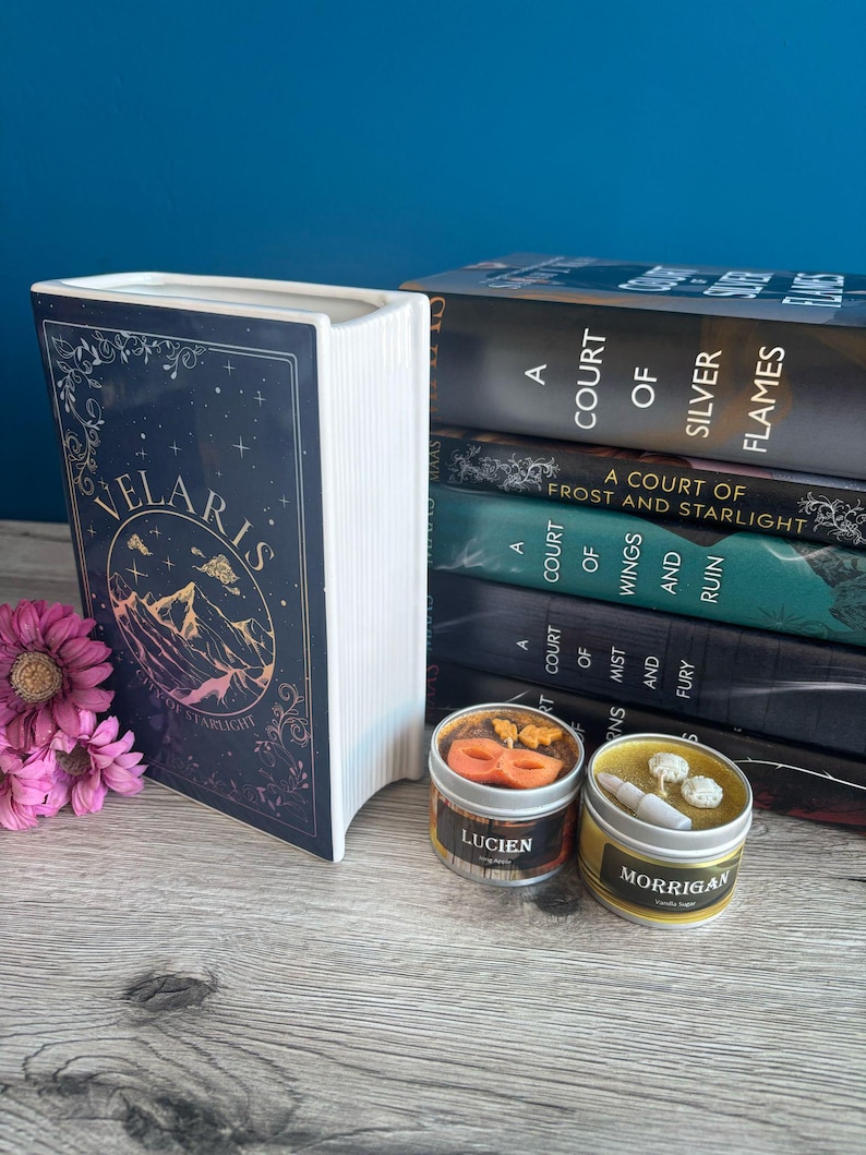 May include: A stack of five books with the title "A Court of Silver Flames" on top. The books are all part of the same series, "A Court of Thorns and Roses". There are two small candles in front of the books, one with a mask design and the text "Lucien" and the other with a flower design and the text "Morrigan".