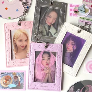 Op de afbeelding: Een verzameling foto-ID-houders in roze, zilver en wit, elk met een stervormig frame en een ketting. De houders tonen foto's van mensen. Decoratieve stickers en cd's met sterbedels zijn ook aanwezig.