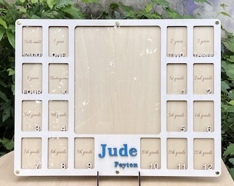 May include: A rectangular light-colored wooden school years photo frame with a large central opening and smaller slots labeled from Hello world to 12th grade. The name Jude Peyton is displayed in blue 3D lettering at the bottom center.