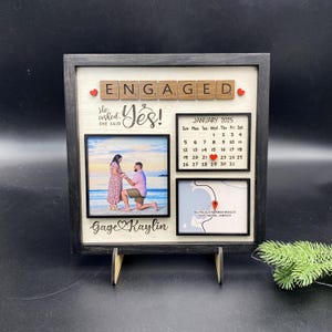 May include: A wooden picture frame with a black border and a white background. The frame has a calendar for January 2025, two photos, and the words "ENGAGED" and "Yes!" in red and black letters. The text "She said" and "He asked" are also included. The names "Gage" and "Kaylin" are written below the photos. The frame is on a wooden stand.