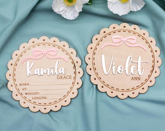 Puede incluir: Dos placas de anuncio de nacimiento de madera con lazos rosas. Una placa dice "Kamila Grace" y tiene espacio para los detalles del nacimiento. La otra placa dice "Violet Ann".