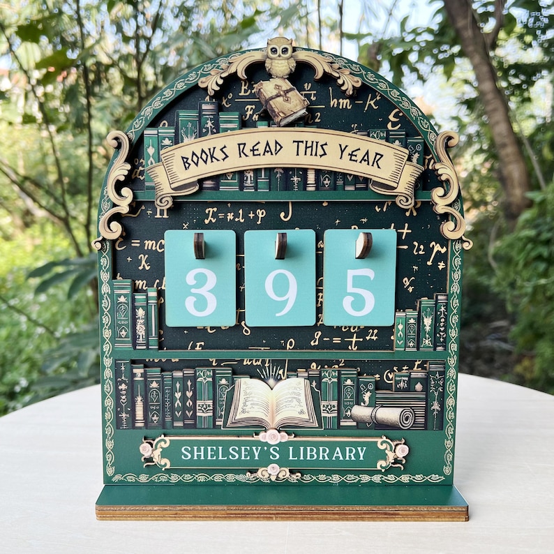 May include: A green wooden bookshelf-themed counter with the text "BOOKS READ THIS YEAR" and the numbers "395" displayed on teal panels. The bottom of the counter reads "SHELSEY'S LIBRARY". An owl and a book are at the top.