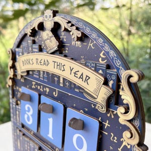 May include: Wooden calendar with a dark blue background and gold accents. The calendar features the text "BOOKS READ THIS YEAR" and displays the numbers 310. An owl and book are at the top.