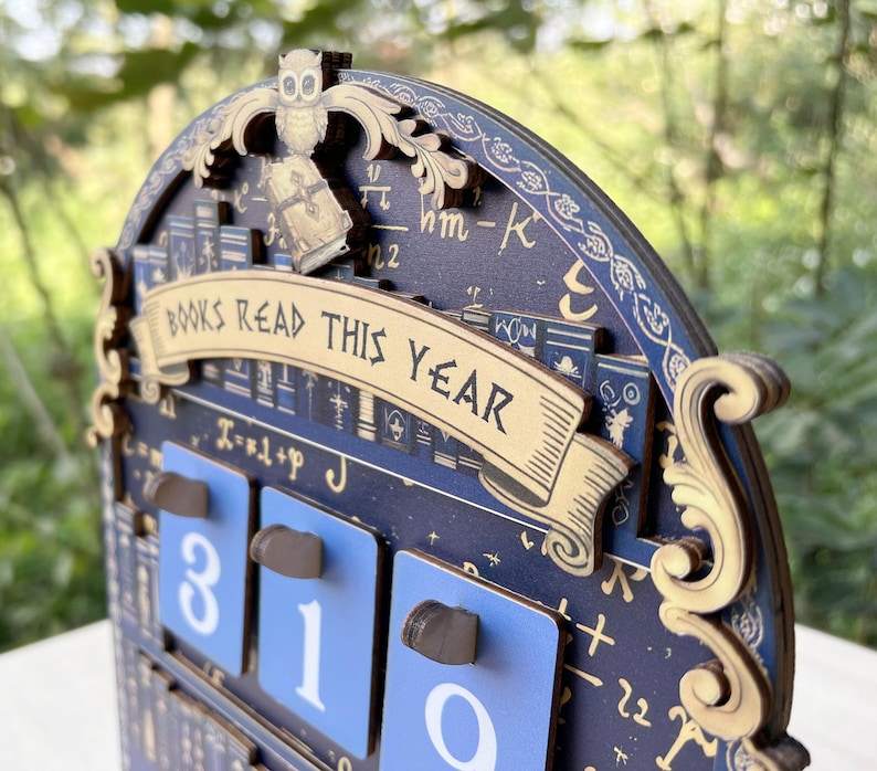 May include: Wooden calendar with a dark blue background and gold accents. The calendar features the text "BOOKS READ THIS YEAR" and displays the numbers 310. An owl and book are at the top.