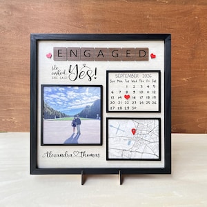 May include: A square black-framed shadow box featuring the word ENGAGED in wooden letter tiles, a photo of a couple, a September 2026 calendar with a red heart on the 15th, a map with a red location pin, and the names Alexandra and Thomas.