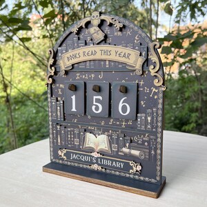 May include: A wooden desk calendar with the text "BOOKS READ THIS YEAR" and the numbers "156" displayed. The calendar has a dark grey background with decorative elements, including an owl, books, and the text "JACQUI'S LIBRARY".