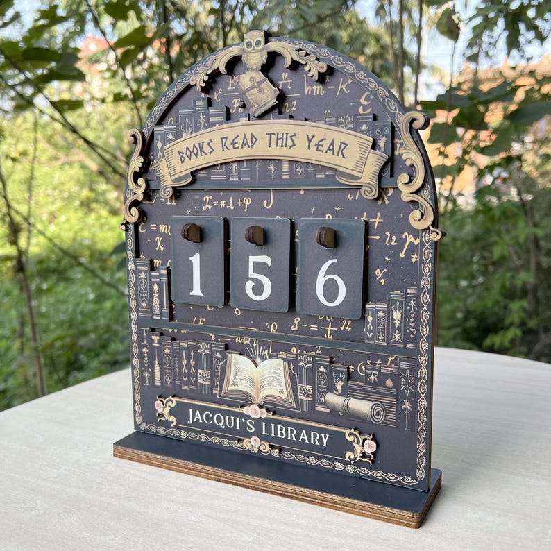 May include: A wooden desk calendar with the text "BOOKS READ THIS YEAR" and the numbers "156" displayed. The calendar has a dark grey background with decorative elements, including an owl, books, and the text "JACQUI'S LIBRARY".