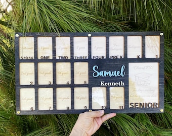 May include: A black wooden photo frame with 12 slots for pictures. The frame has gold lettering that says "Hello World! 5.19.14", "One", "Two", "Three", "Four", "Pre K", "K", "1st Grade", "2nd Grade", "3rd Grade", "4th Grade", "5th Grade", "6th Grade", "7th Grade", "8th Grade", "9th Grade", "10th Grade", "11th Grade", and "Senior". The frame also has a space for a name and the text "Class of 2022". The name "Samuel Kenneth" is written in blue cursive lettering.