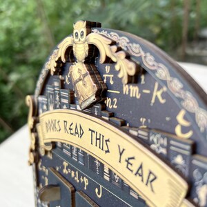 May include: A wooden book tracker with an owl and book design. The words "BOOKS READ THIS YEAR" are displayed on a banner. The tracker is dark blue with gold accents and features book illustrations and mathematical symbols.