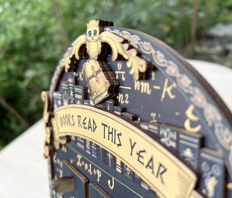 May include: A wooden book tracker with an owl and book design. The words "BOOKS READ THIS YEAR" are displayed on a banner. The tracker is dark blue with gold accents and features book illustrations and mathematical symbols.