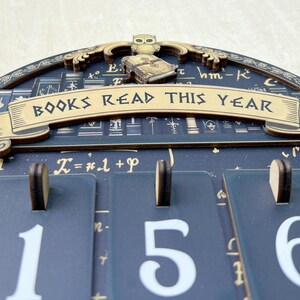 May include: A wooden wall calendar with the text "BOOKS READ THIS YEAR". The calendar has a dark blue background with gold accents and a decorative owl and book on top. The numbers 1, 5, and 6 are displayed.