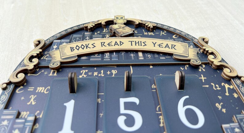 May include: A wooden wall calendar with the text "BOOKS READ THIS YEAR". The calendar has a dark blue background with gold accents and a decorative owl and book on top. The numbers 1, 5, and 6 are displayed.