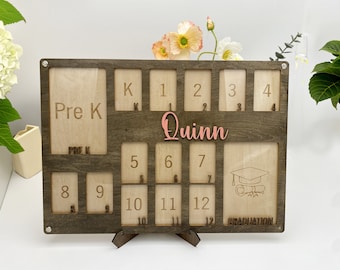 May include: A wooden photo frame with 12 slots for pictures, labeled with the grades Pre-K through 12. The frame is engraved with the name "Quinn" and the word "Graduation".