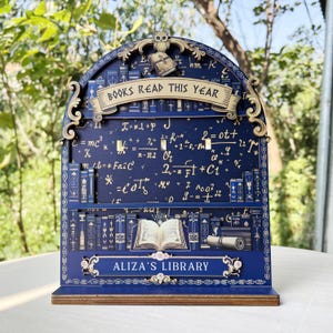May include: A dark blue wooden bookshelf-shaped display with the text "BOOKS READ THIS YEAR" and "ALIZA'S LIBRARY". The display features an owl, equations, and illustrations of books. The shelf is made of wood and has a decorative design.