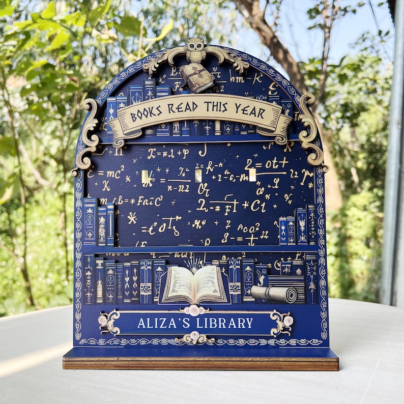 May include: A dark blue wooden bookshelf-shaped display with the text "BOOKS READ THIS YEAR" and "ALIZA'S LIBRARY". The display features an owl, equations, and illustrations of books. The shelf is made of wood and has a decorative design.