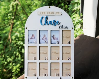 May include: An arched white wooden photo display board featuring the text "ONE YEAR OF Chase Mae" in blue and black lettering. The board contains twelve rectangular slots for photos, labelled with numbers 1 through 12 and corresponding month markers.