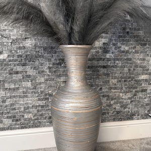 May include: A decorative silver vase with a textured, ridged design, filled with gray feathery plumes. The vase is placed against a gray brick wall. The vase is approximately 60 cm tall.