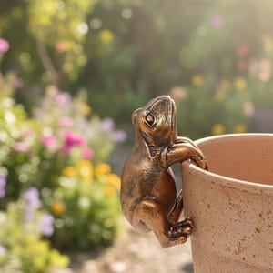 Könnte beinhalten: Eine bronzefarbene Froschfigur, die sich am Rand eines Blumentopfs aus Terrakotta festhält. Der Frosch ist mit strukturierter Haut detailliert und vor einem verschwommenen Hintergrund aus bunten Blumen und Grün angeordnet.