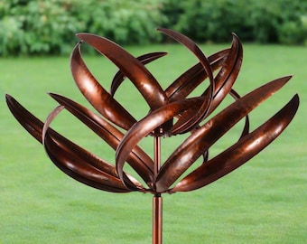 Op de afbeelding: Een koperkleurige metalen tuinsculptuur met een bloemachtig ontwerp. De sculptuur heeft gebogen, bladvormige elementen die rond een centrale steel zijn gerangschikt. De sculptuur staat tegen een achtergrond van groen gras en gebladerte.