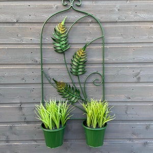 May include: A green metal wall planter with two green pots and green plants. The planter features a decorative fern design.