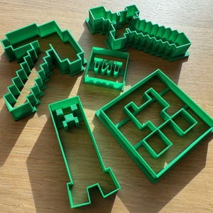 Könnte beinhalten: Ein Set aus fünf grünen Plastik-Ausstechern in Form von Minecraft-Gegenständen. Die Ausstecher beinhalten eine Spitzhacke, ein Schwert, einen Block, einen Creeper und das Wort "TNT".