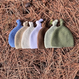 Puede incluir: Una colección de siete gorros de ganchillo dispuestos en fila, con un diseño de nudo en la parte superior. Los gorros se presentan en una variedad de colores sólidos, incluyendo azul, beige, crema, blanco, morado claro, marrón, verde salvia y oliva.