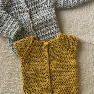 May include: Two handmade crochet baby sweaters. One is a long-sleeved gray cardigan with wooden buttons. The other is a sleeveless mustard yellow vest with wooden buttons. Both are made with a textured stitch.
