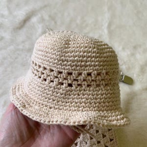 May include: A crocheted bucket hat in a light beige colour. The hat has a wide brim and a crocheted band around the crown. The hat is made with a simple stitch pattern.