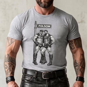 May include: Light grey t-shirt with a graphic of two men in leather clothing, standing under a street sign that says "FOLSOM". The shirt is a sport grey colour.