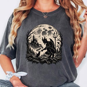 May include: A charcoal grey short-sleeve t-shirt featuring a graphic print of a horned devil figure touching a cat on the nose with the text BOOP. The scene includes a large full moon, a castle on a cliff, and a forest silhouette in black ink.