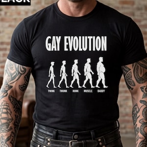 May include: A black short-sleeved t-shirt featuring the white text GAY EVOLUTION above a graphic showing five human silhouettes labelled TWINK, TWUNK, HUNK, MUSCLE, and DADDY. The word BLACK is printed in the upper left corner of the image.