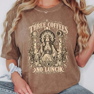 May include: An espresso-coloured short-sleeve t-shirt featuring a graphic print of a seated figure holding a cup, surrounded by pillars, celestial symbols, and coffee-themed elements. The text reads THREE COFFEES NO LUNCH in a decorative font.