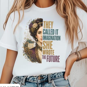 May include: A white t-shirt featuring a graphic print of a woman with an elaborate floral hairstyle and mechanical gear accents. The text reads, "THEY CALLED IT IMAGINATION SHE WROTE THE FUTURE" in stacked, multi-coloured lettering next to the portrait.