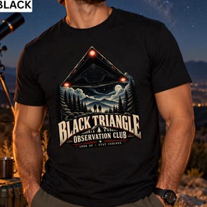 May include: A black t-shirt featuring a graphic of a triangular UFO hovering over a forest and mountains at night. The text reads BLACK TRIANGLE OBSERVATION CLUB and LOOK UP STAY CURIOUS. The design includes two silhouettes of people standing in the woods.