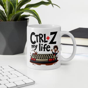 Può includere: Tazza in ceramica bianca con un'illustrazione in bianco e nero di una tastiera con una persona che ci cade dentro. Il testo "CTRL+Z my life" è stampato sulla tazza.