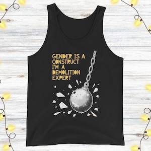 Könnte beinhalten: Schwarzes Tanktop mit dem Text "GENDER IS A CONSTRUCT I'M A DEMOLITION EXPERT" in goldfarbenen Buchstaben. Eine silberne Abrissbirne ist abgebildet, die schwingt und eine Oberfläche zerbricht. Das Design ist mittig auf dem Tanktop platziert.