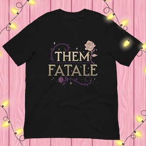 May include: Black t-shirt featuring the text "THEM FATALE" in a vintage style, with rose illustrations. The design includes a light purple floral border and a light pink rose. The shirt has a classic crew neck.