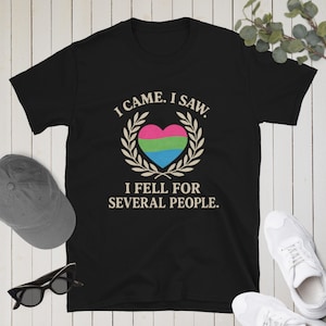Peut inclure: T-shirt noir avec l'inscription "I came. I saw. I fell for several people." Le motif comprend un cœur rose, vert et bleu, entouré d'une couronne de laurier. Le t-shirt est à col rond et à manches courtes.