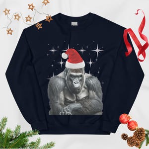 Könnte beinhalten: Ein dunkelblaues Sweatshirt mit Rundhalsausschnitt und einer Grafik eines grauen Gorillas, der eine rot-weiße Weihnachtsmütze trägt. Der Gorilla ist mittig auf der Brust platziert und von weißen Sternen auf dem dunklen Stoff umgeben.